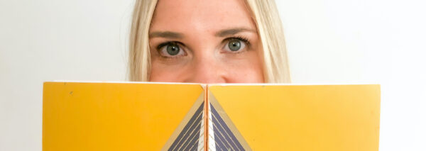 A person with blonde hair peers over a closed yellow book with a geometric design against a plain white background.