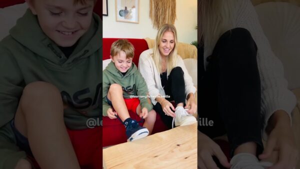 A person and child smile while tying shoelaces on a red couch. Framed wall art and plant decor are visible in the background.