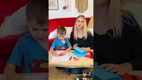 A child and a person sit at a table doing crafts with colored paper, crayons, and scissors in a cozy living room.