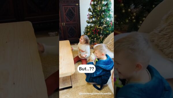Two children sitting near a table with a Christmas tree in the background, reacting with surprise. Holiday decorations and a wooden cabinet present.