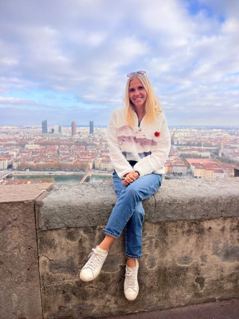 A person sits on a stone wall overlooking the city of Lyon, with tall modern buildings in the background under a cloudy sky.