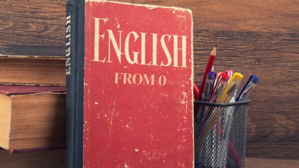 Red book titled "English From 0" stands upright, alongside a metal holder containing colorful pens, against a wooden background.
