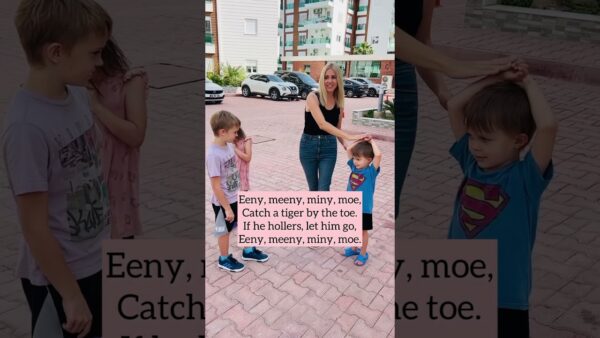 A person and three children play "Eeny, Meeny, Miny, Moe" in a brick courtyard with parked cars and apartment buildings in the background.