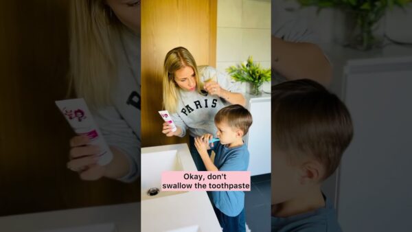 Camille helps Kairo with toothpaste in a bathroom. Kairo holds a brush, standing by a sink with flowers nearby. - Teach your child how to brush their teeth in English