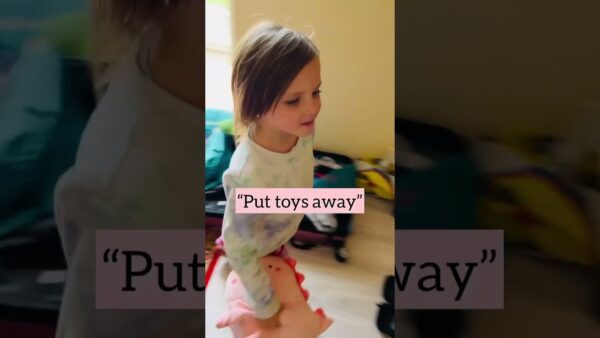 A child holds a pink stuffed toy indoors with text overlay saying, "Put toys away," suggesting tidying up.