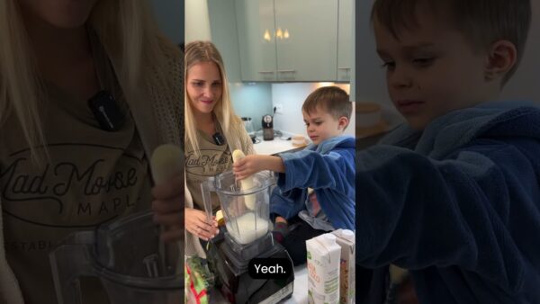 A person and child prepare a smoothie in a kitchen, using a blender. The child adds a banana to the blender.
