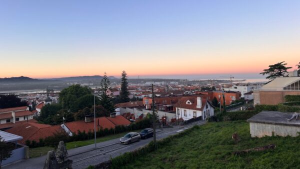 Sunset view over a hillside town with red-roofed buildings, trees, and distant cranes. Peaceful evening sky transitions from orange to blue.