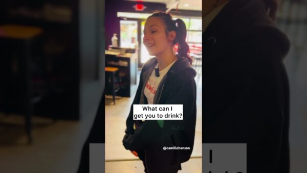 A person in a dimly-lit cafe, wearing casual clothing, smiles while asking, “What can I get you to drink?” No landmarks visible.