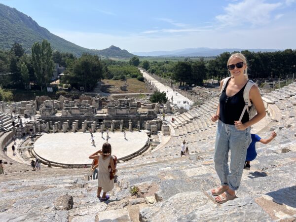 A person stands in the ancient Greek Theatre of Ephesus, with ruins and a scenic mountainous background under a clear sky.