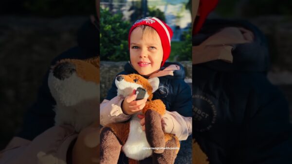 A child wearing a red hat holds a plush toy outdoors, with greenery in the background.