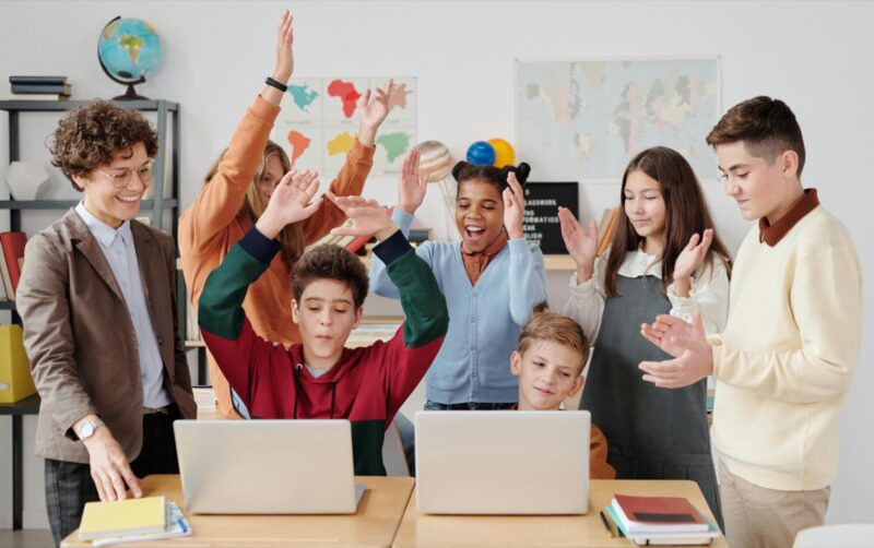 A person and six children celebrate in a classroom, with two laptops open. Maps and a globe are visible in the background.