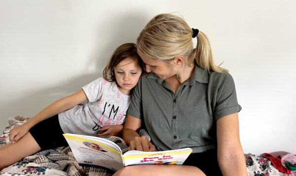 A person and child sit closely on a bed, sharing a book in a warm, intimate setting with a plain white background.