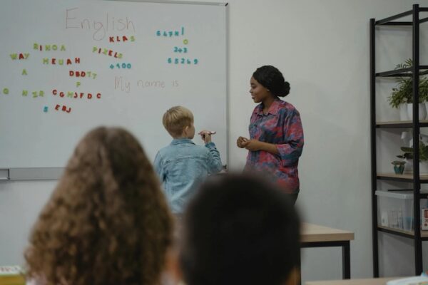 A person teaches a child English using a whiteboard in a classroom, with colorful magnetic letters and numbers, alongside blurred students.