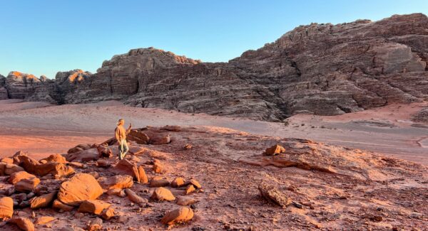 A person stands on rocky terrain in Wadi Rum desert, Jordan, surrounded by dramatic red sandstone formations under a clear blue sky.